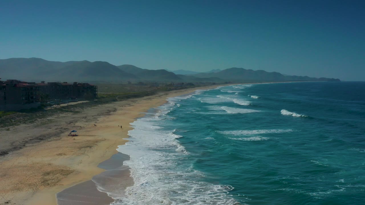 A high oblique view of Cerritos shoreline with turquoise water and foamy breaks on the right, pale sand on the left, low vegetation near the buildings, mountain horizon, and no visible people.