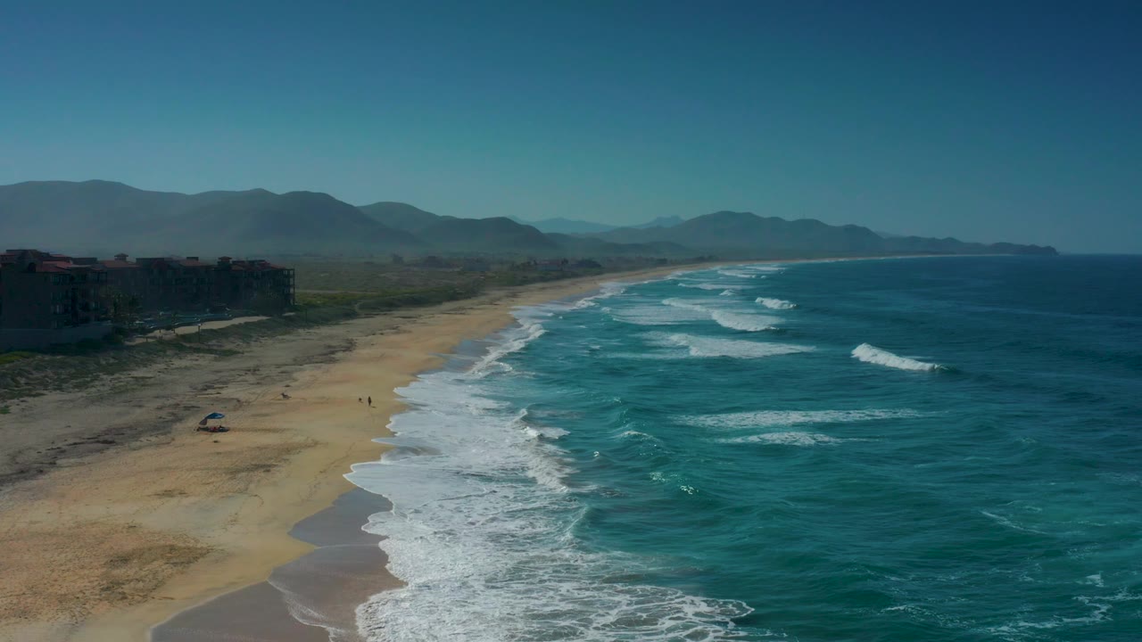 Cerritos shoreline seen from above, with a small shade object or umbrella-like shape on the sand, layered white waves, dark buildings at left, distant mountains, and no visible people.