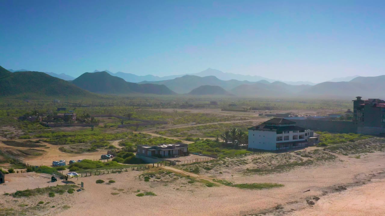 Opening aerial frame over Cerritos, Baja California Sur, showing sandy foreground, scattered green shrubs, a white building, dirt roads, distant mountains, and no visible people.