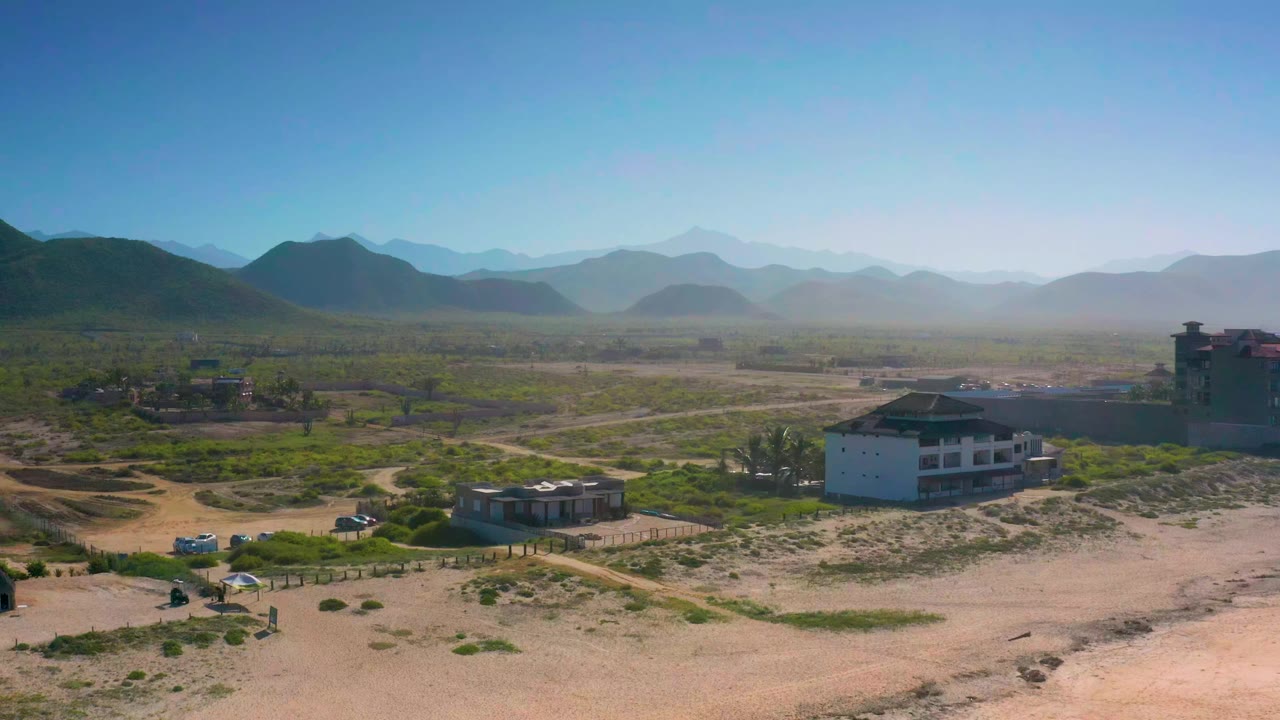Aerial frame in Cerritos, Baja California Sur, with dirt tracks crossing the valley, cars near the lower left, low buildings in the midground, mountains beyond, and no visible people.