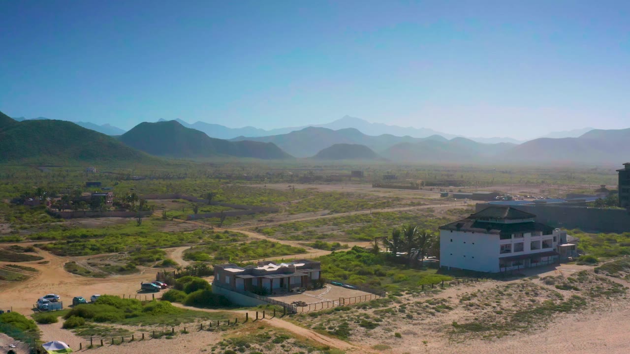 Aerial frame over Cerritos, Baja California Sur with a white multi-story building, smaller houses, parked vehicles, dry scrub, mountain ridges, and no visible people.