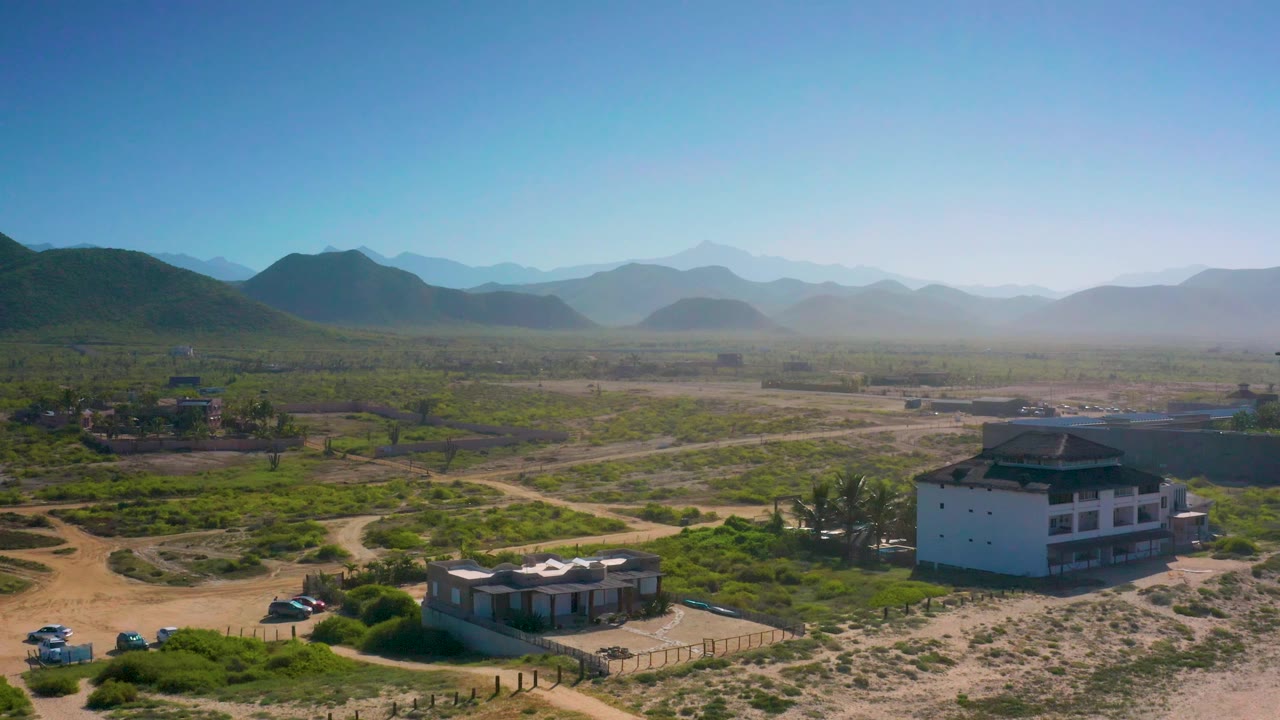 Wide aerial view in Cerritos, Baja California Sur with houses along sandy roads, green scrub patches, a hazy mountain background, and no visible people.