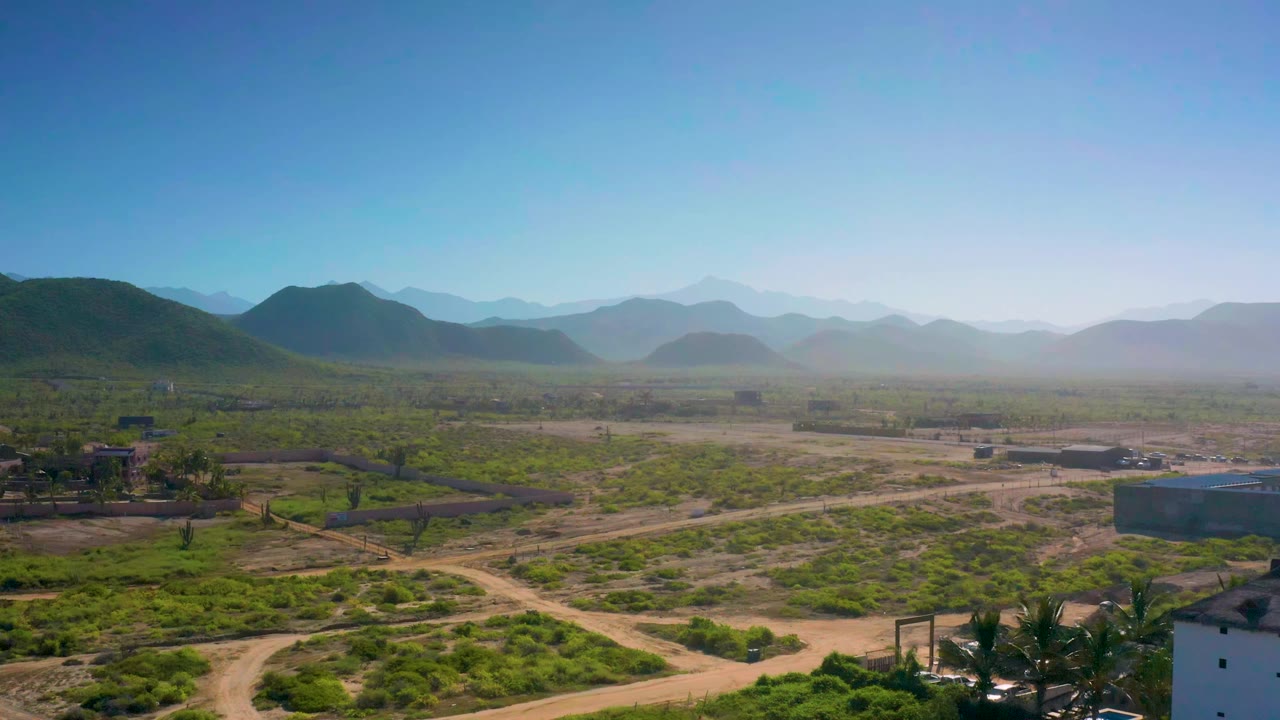 Elevated Cerritos landscape frame showing intersecting dirt tracks through green bushes, small structures in the midground, hazy mountain layers, blue sky, and no visible people.