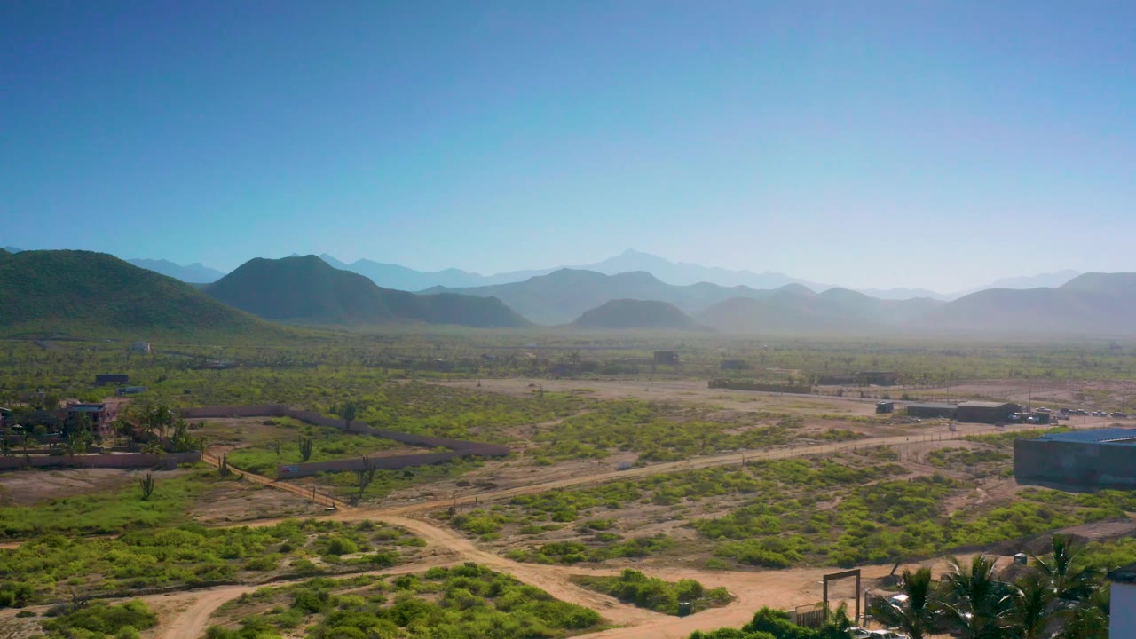 A wide aerial view near Cerritos with a low rectangular building on the right, palms near the lower edge, open scrubland, distant hills, bright sky, and no visible people.
