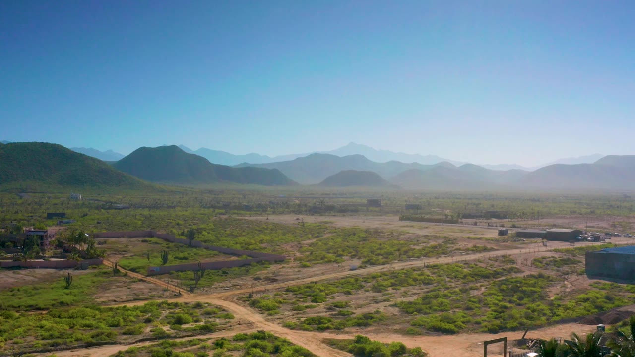 Elevated frame over Cerritos scrubland with dirt roads running across the foreground, low buildings in the midground, a hazy hill range beyond, and no visible people.