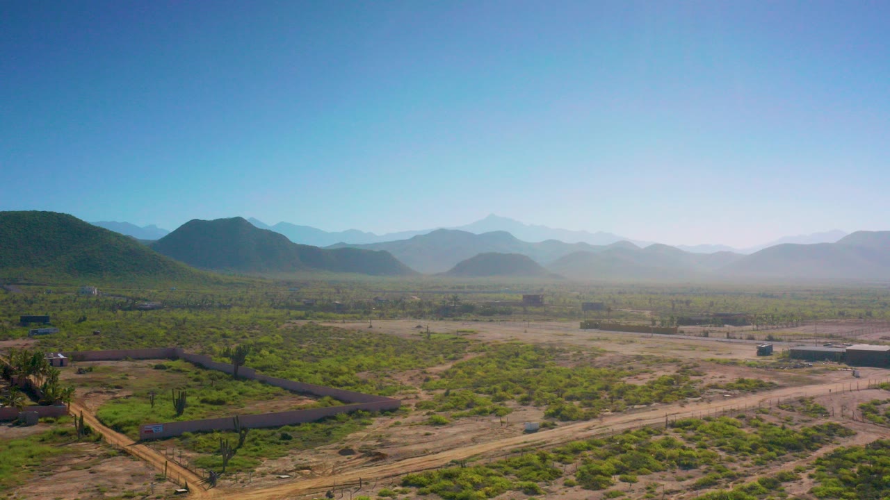 Opening aerial frame over Cerritos, Baja California Sur scrubland with green bushes, dirt tracks, small structures, hazy mountain ridges, and no visible people.