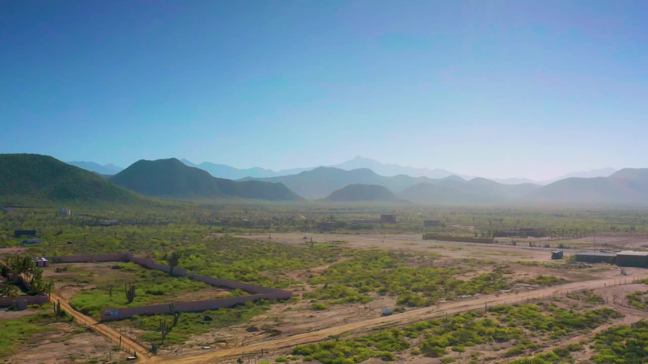 Wide aerial landscape with low green vegetation, dirt tracks, and layered mountains.