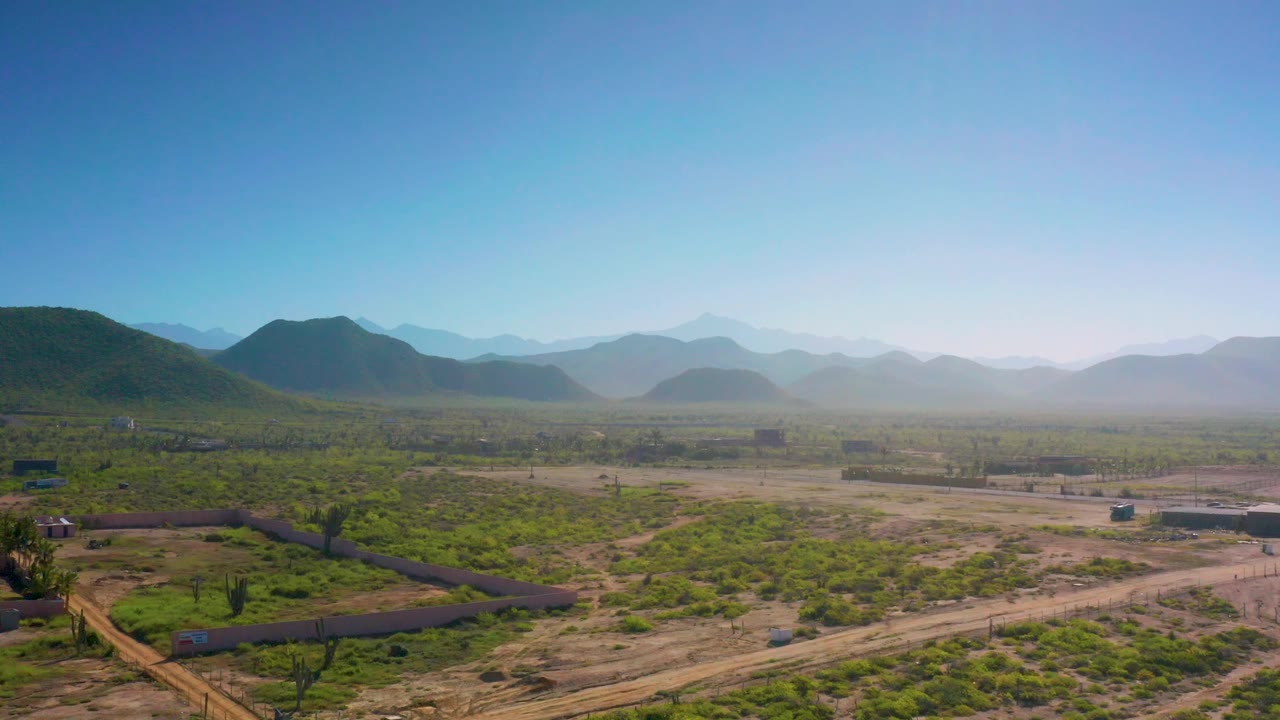 Cerritos aerial frame with a greener foreground, pale dirt roads, a few walled areas, mountain silhouettes in the distance, clear daylight, and no visible people.
