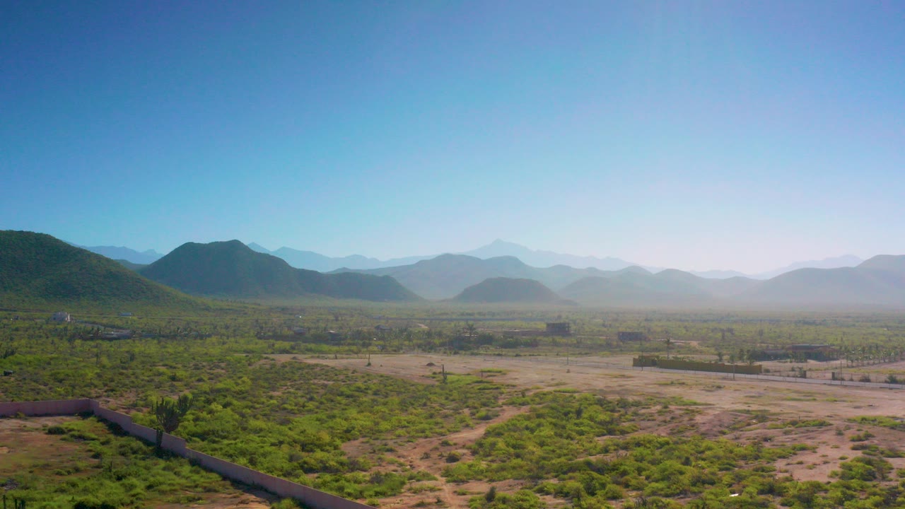 Wide aerial frame over Cerritos, Baja California Sur, with green desert scrub, sandy ground, distant mountain ridges, pale morning haze, and no visible people.