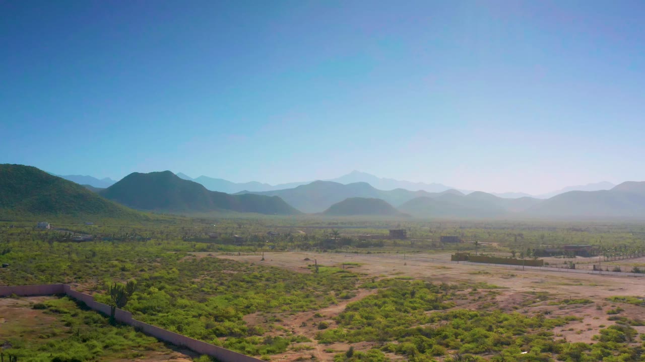 Drone-height view across Cerritos desert terrain with low brush in front, open sandy flats, layered blue mountains in the distance, and no visible people.