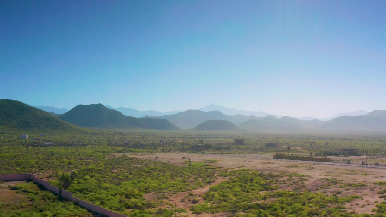 Cerritos aerial view showing scrub-covered desert, pale dirt areas, low walls or fences, small distant structures, hazy mountain layers, and no visible people.