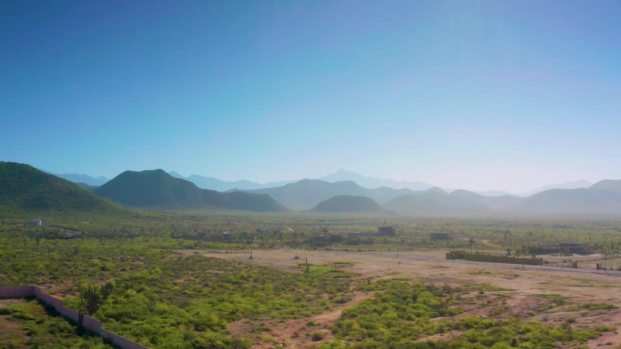 Aerial frame over Cerritos with dense green scrub in the foreground, sandy open ground through the center, hazy mountains beyond, and no visible people.