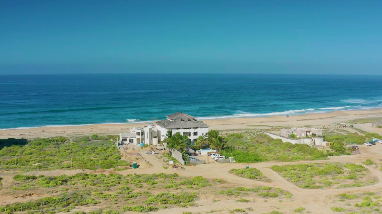 Cerritos aerial frame with the large white house slightly left of center, scrubby beach vegetation below, a sandy beach beyond, and no visible people.