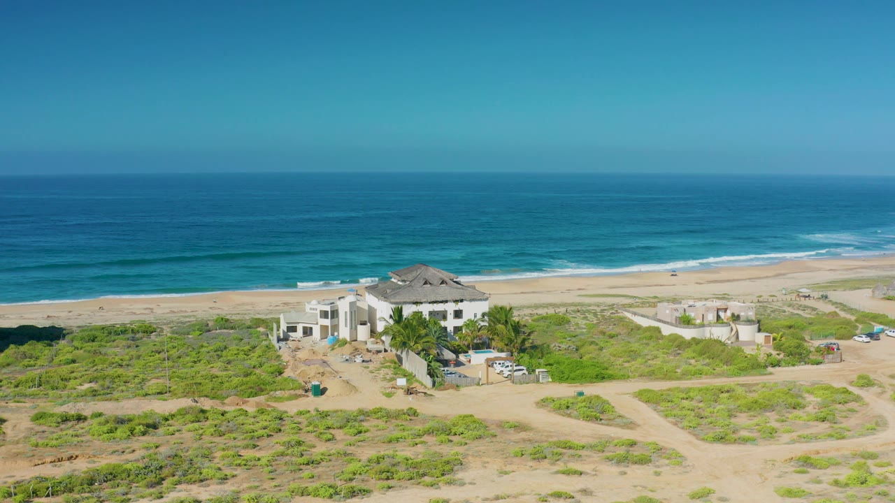 A high coastal view in Cerritos shows the beachfront house near the lower center, pale sand, low green growth, turquoise water bands, and no visible people.