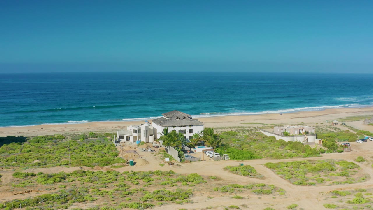 Aerial beach scene with a white house, dune vegetation, and calm blue water.
