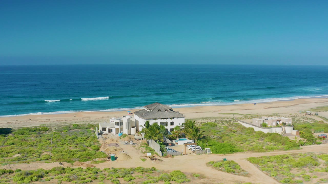 Aerial opening frame over Cerritos, Baja California Sur, showing a sand-colored mansion below the beach, blue ocean beyond, low green scrub in front, and no visible people.