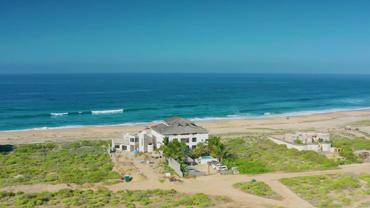 Aerial frame in Cerritos with the mansion centered lower in view, palm trees and a blue pool near the house, surf bands on the beach, and no visible people.