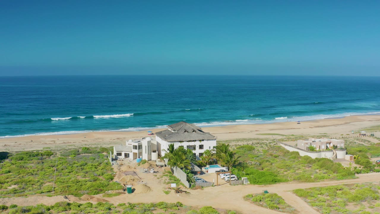 Aerial view over Cerritos showing the beach house slightly lower in frame, surrounding dirt roads and scrub, a long sand strip, breaking waves, and no visible people.