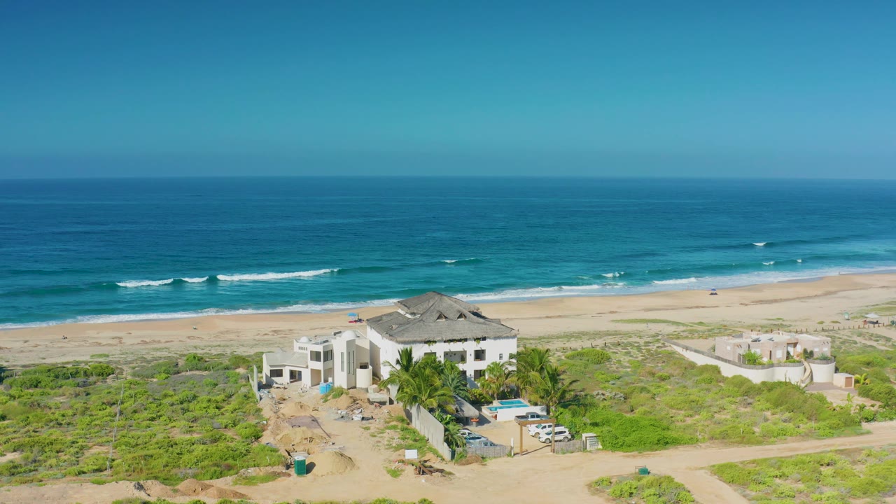 Wide aerial coastal view with a mansion, beach, surf, and clear blue sky.