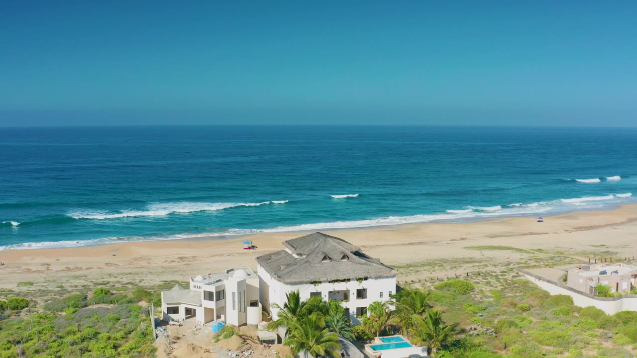 Aerial frame over Cerritos, Baja California Sur showing a white beachfront house, low dune vegetation, pale sand, turquoise surf, blue sky, and no visible people.