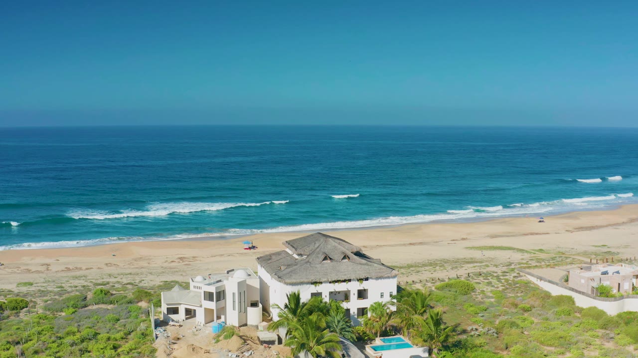 Cerritos aerial view with the white house and palms lower in frame, a small umbrella on the sand, breaking waves offshore, and no visible people.