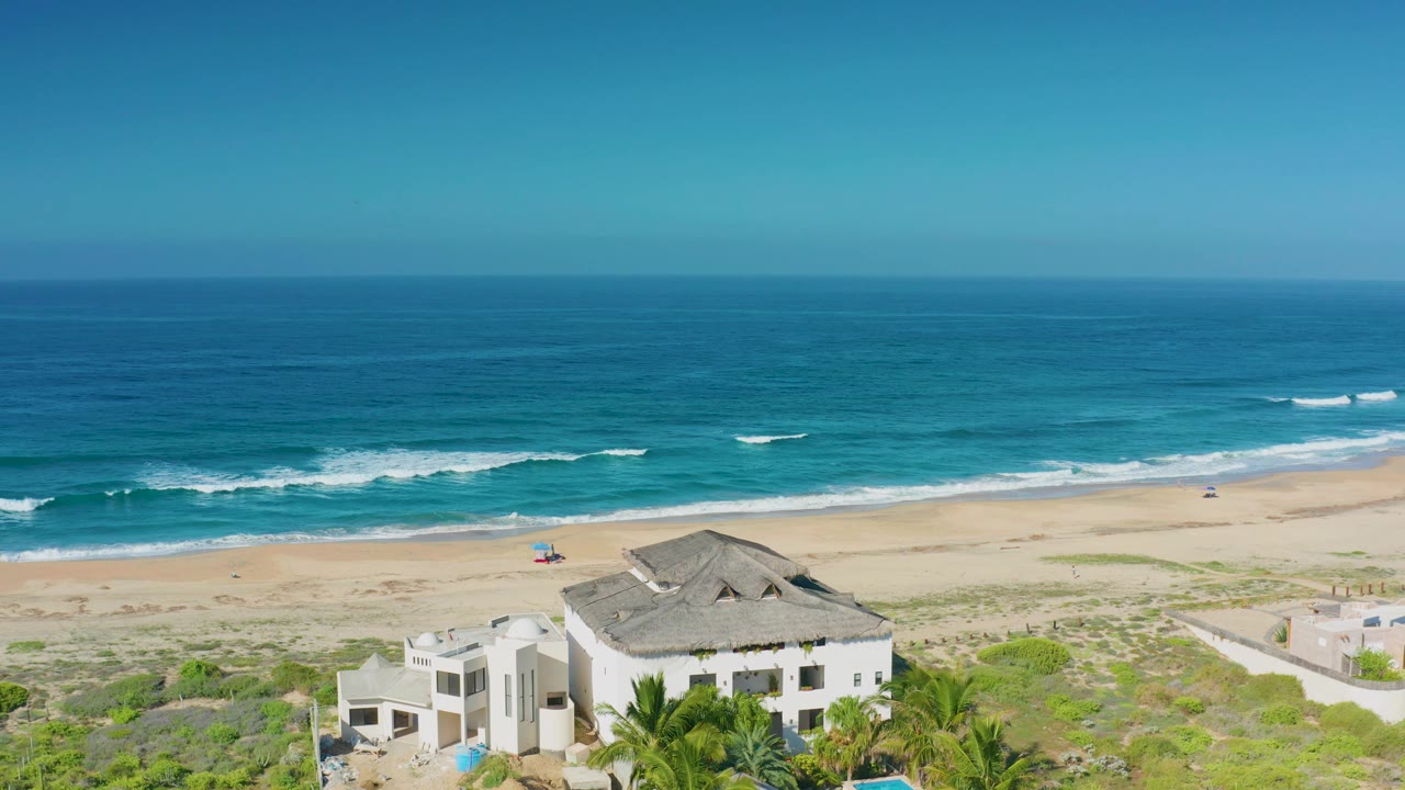 Aerial view in Cerritos with the large white house partly lower frame, scrubby dunes, pale beach, layered surf, blue ocean, and no visible people.