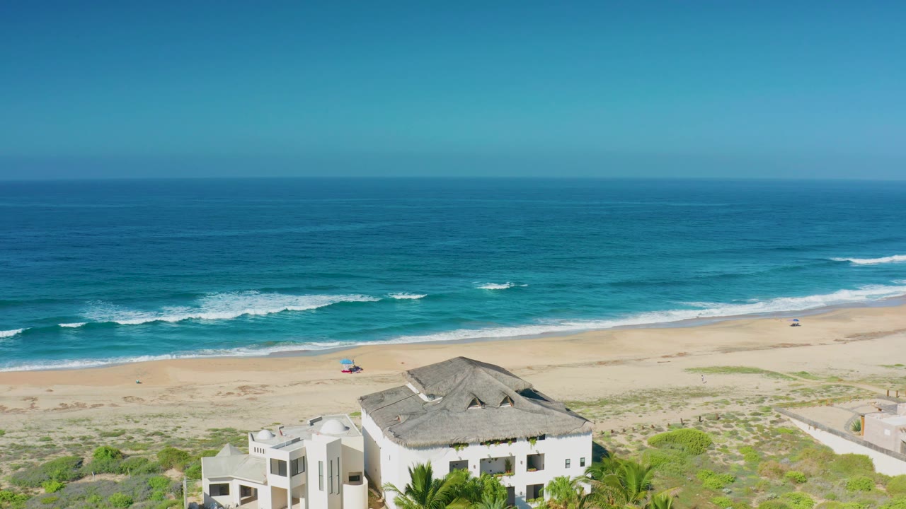 Aerial coastline view with white house, sandy beach, small umbrella, surf, and horizon.