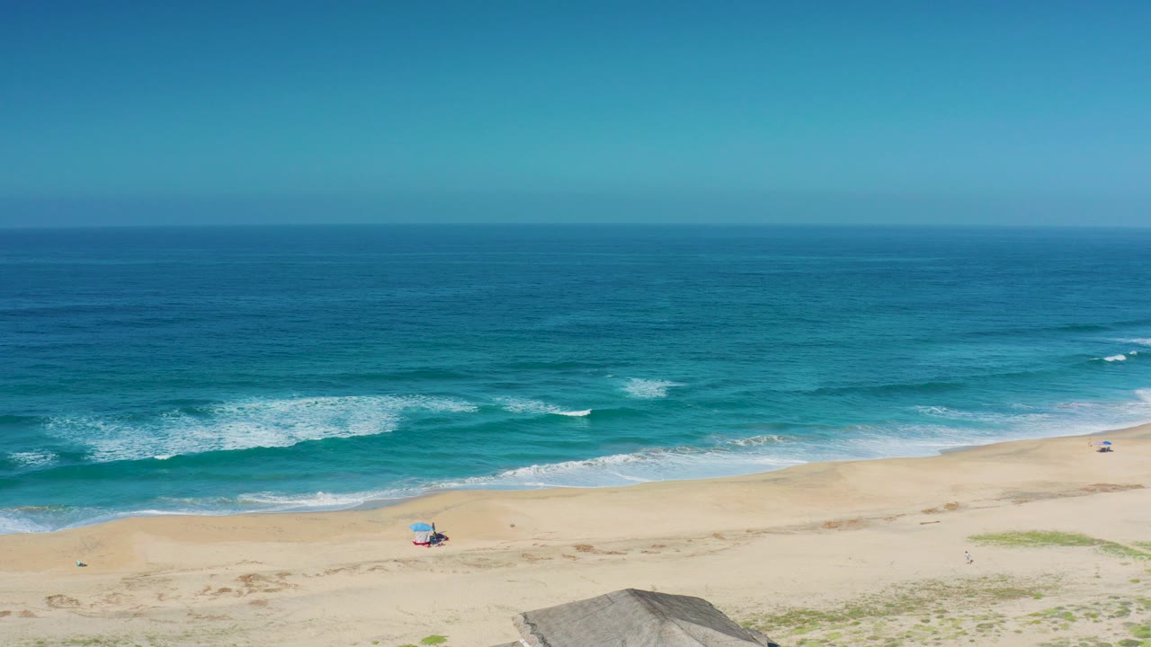 Wide aerial-style view of Cerritos beach with turquoise waves breaking toward pale sand, a small colorful chair setup near the lower center, and no visible people.