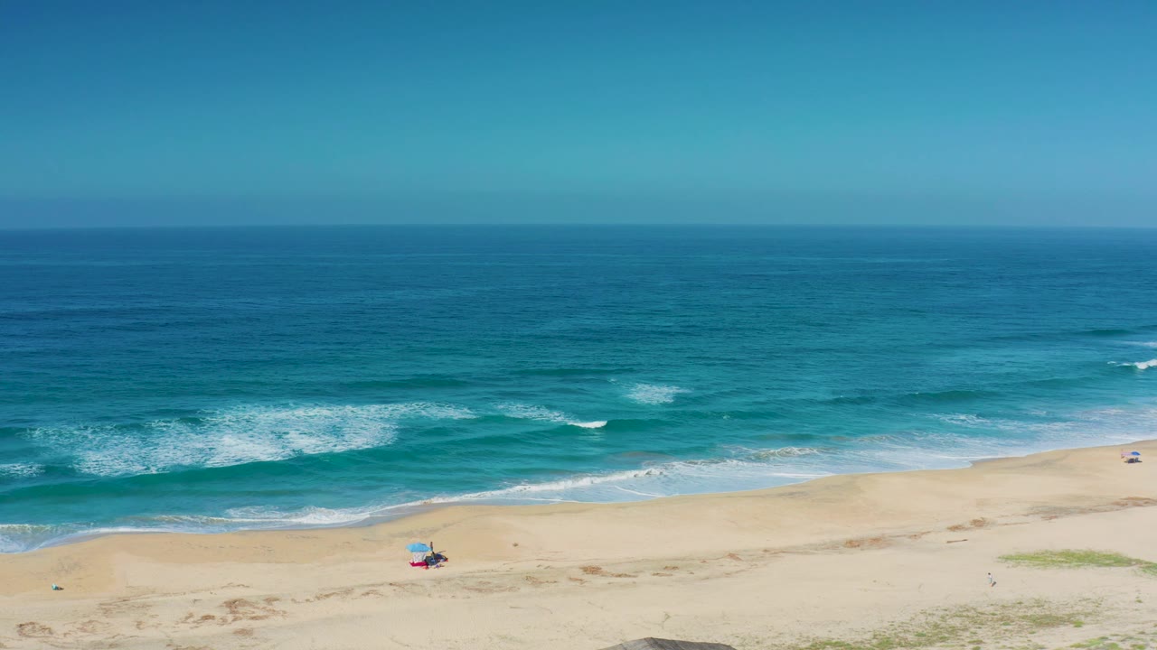 A bright Cerritos beach frame with a level horizon, turquoise ocean, white breaking waves, pale sand, a tiny colorful beach chair setup, and no visible people.