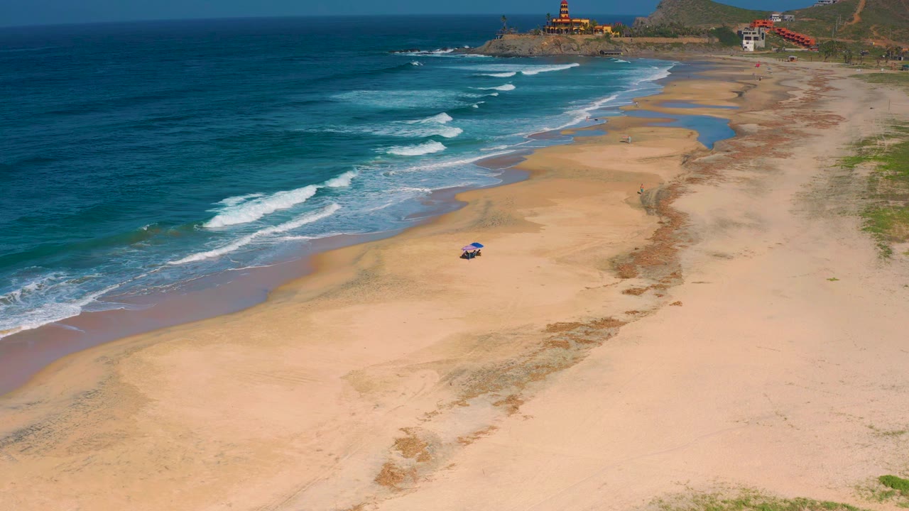 Aerial frame over Cerritos with curved white waves, pale sand, shallow wet channels near the shoreline, distant buildings on the point, and no visible people.