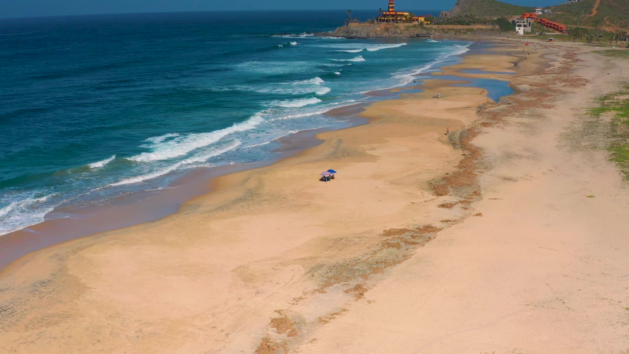 Drone view of Cerritos beach with deeper blue water at left, staggered white wave breaks, open sand through the center, coastal buildings in the distance, and no visible people.