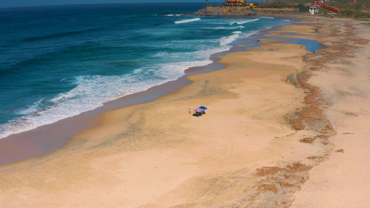 Cerritos beach aerial at 0:01 shows the surf band slightly farther down the shore, with open sand, tidal wet patches, and a small blue umbrella setup below center.