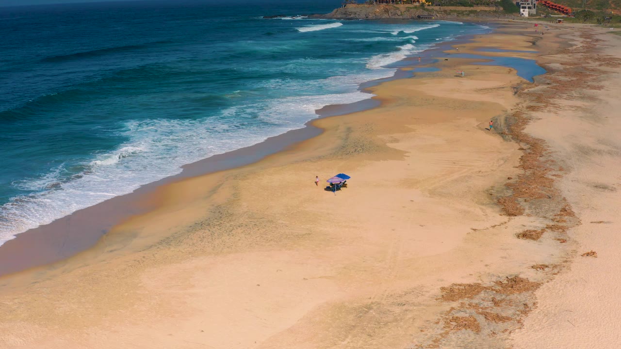 At 0:02, the Cerritos shoreline fills the frame with stronger white foam at the waterline, broad dry sand, a blue umbrella setup, and no recognizable people visible.