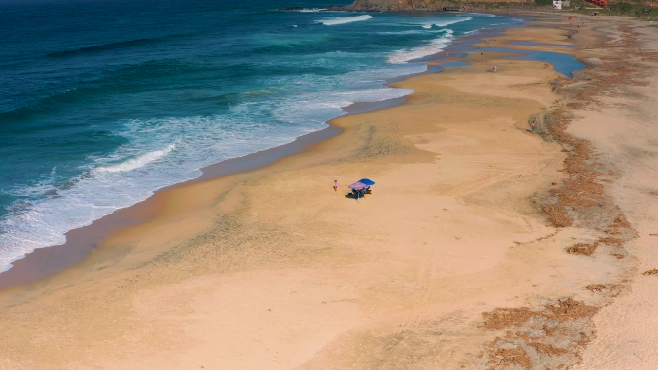 Blue ocean waves break beside a mostly empty beach with a small umbrella setup on the sand.