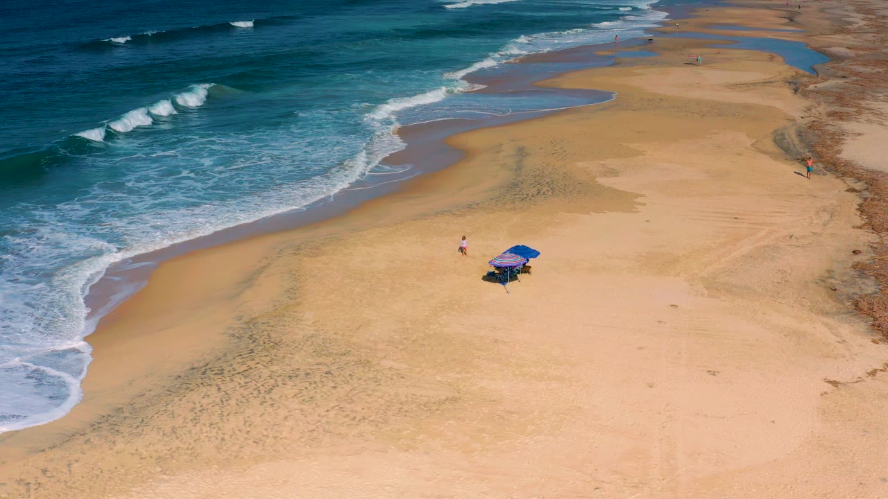 Aerial view of Cerritos beach with teal surf on the left, broad sand in the center, two blue umbrellas near the lower right, and small visible people nearby.