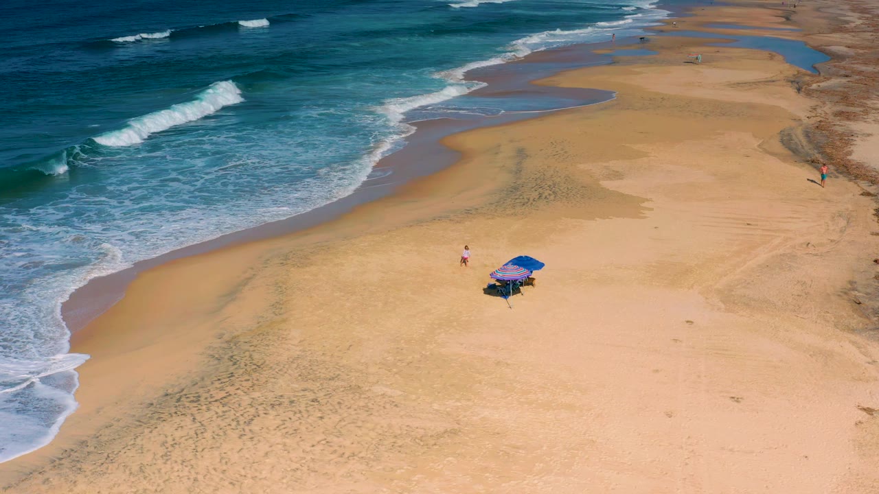 A high diagonal view of Cerritos beach shows foamy surf reaching the left edge, pale sand across most of the frame, two blue umbrellas, and tiny visible beachgoers.