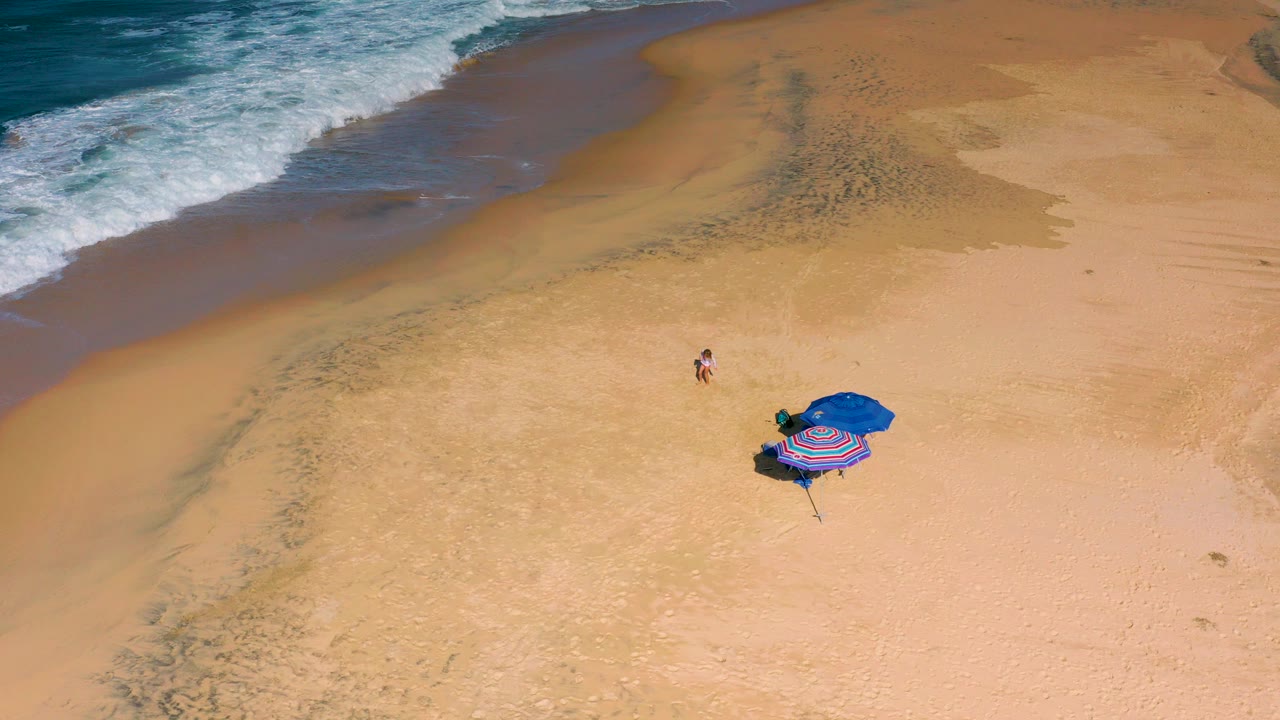Overhead view of Cerritos beach in Baja California Sur, Mexico, with surf at the top, open sand below, a child near center, and blue and striped umbrellas lower right.