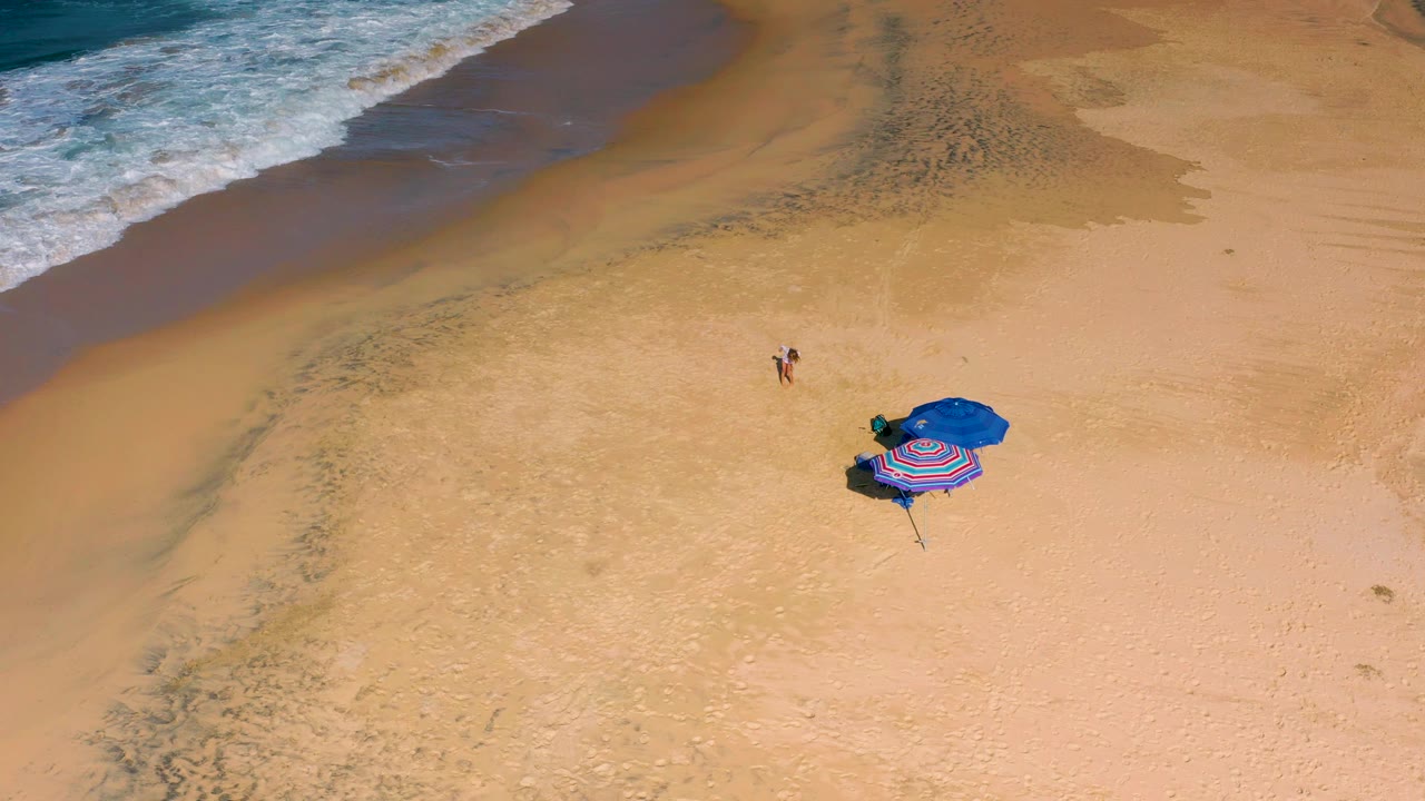 Overhead Cerritos beach view with the child slightly closer to the umbrellas, surf curling along the top left, and broad textured sand filling the frame.