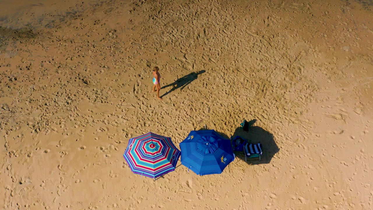 Top-down view of Cerritos beach sand with two colorful umbrellas near the lower frame and a woman walking across the upper sand.