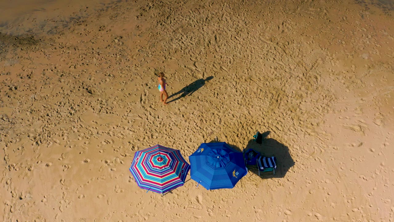 The woman remains in the upper sand area of the Cerritos beach frame, with the patterned umbrella and blue umbrella grouped below her.