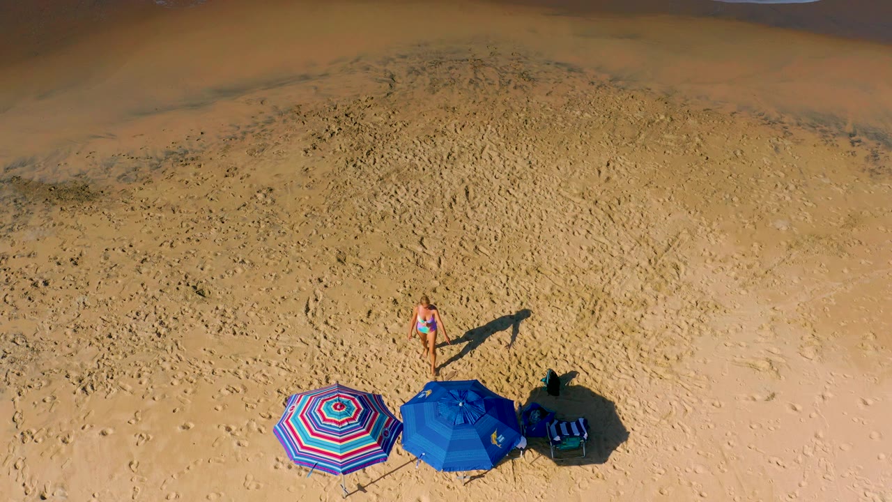 Overhead Los Cabos beach frame with the woman closer to the umbrella cluster, a narrow strip of ocean foam near the top right, and strong sun casting defined shadows on the sand.