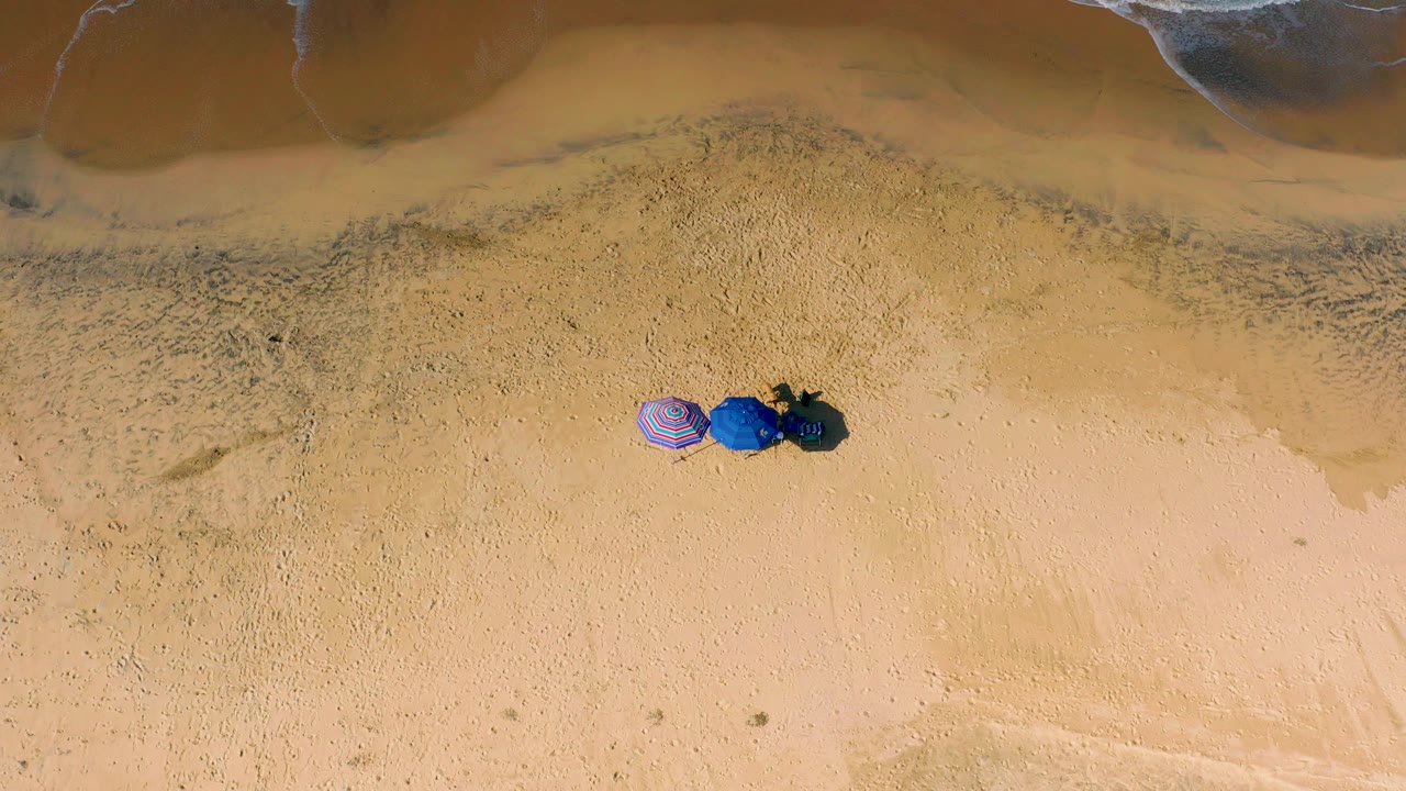 Overhead view at Cerritos showing a small striped umbrella, blue beach fabric, and dark bags on tan sand, with a mottled wet-sand band and surf along the top, with no visible people.