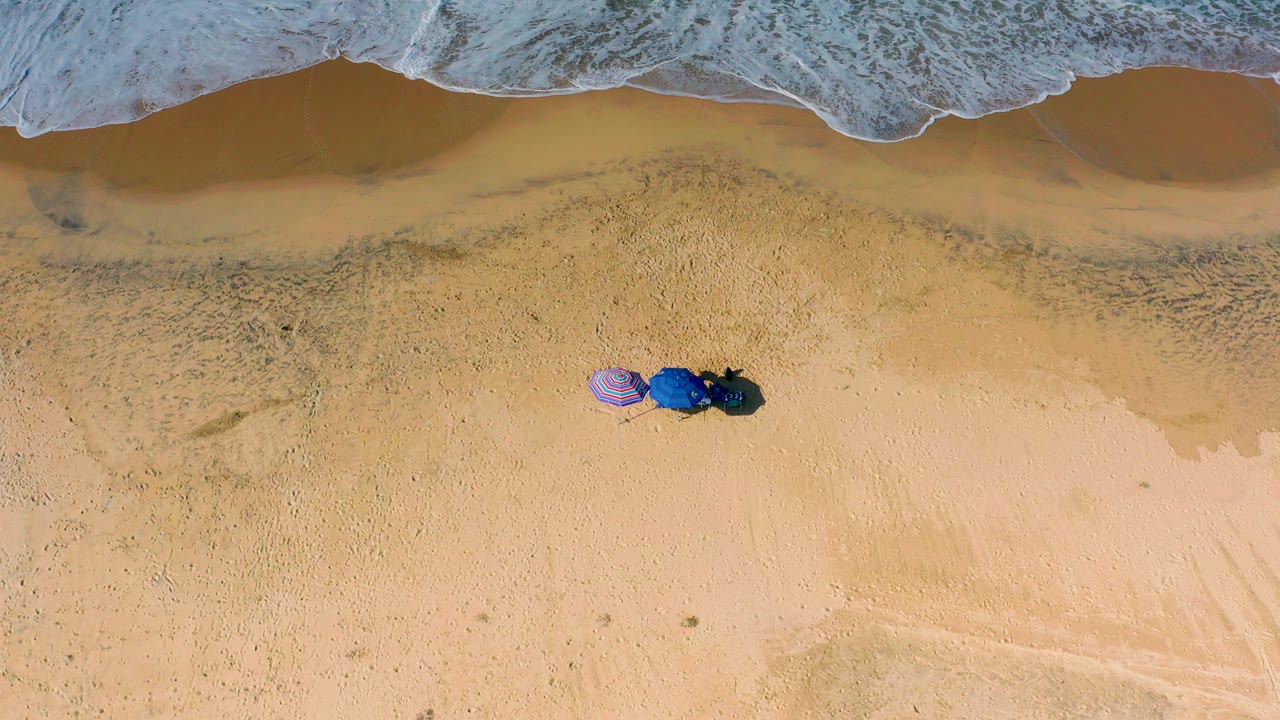 Overhead view of Cerritos beach in Baja California Sur with foamy surf at the top, open sand below, two umbrellas and dark beach gear near the waterline, and no visible people.