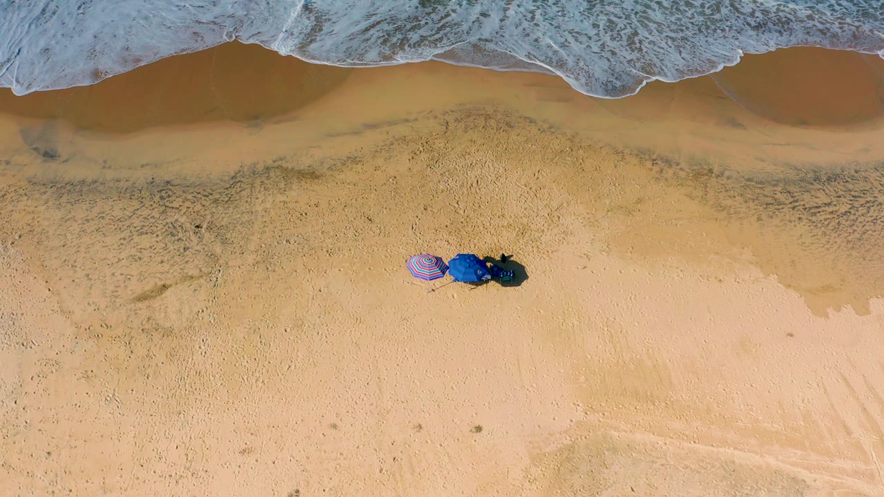 Top-down frame of Cerritos beach showing a band of white surf along the upper edge, tan wet sand, the umbrellas and gear slightly left of center, and no visible people.