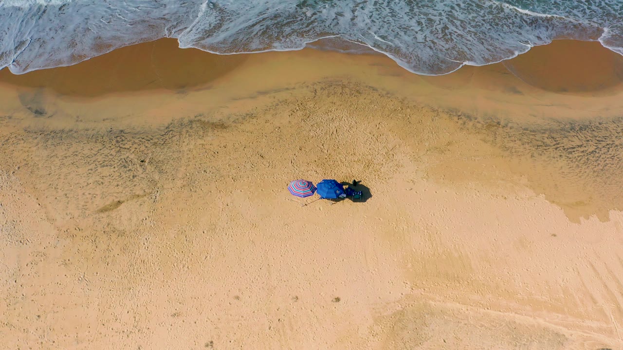 Drone-style overhead view in Cerritos with textured foam pushing across the top of frame, broad sand below, umbrellas and dark gear near center, and no visible people.