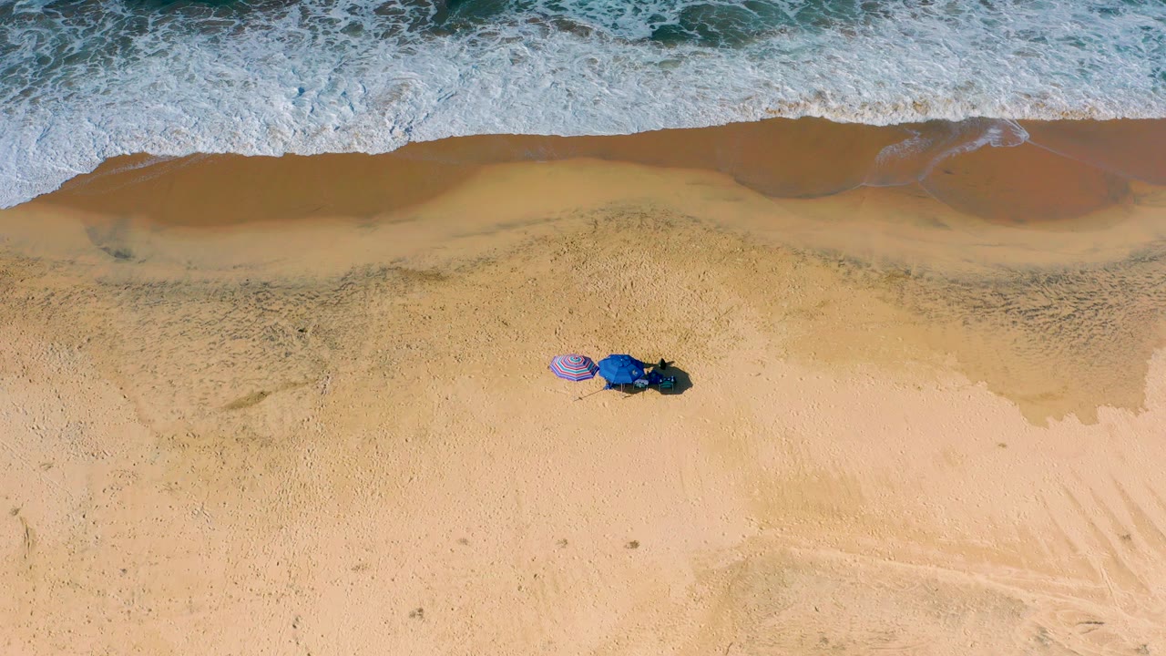Overhead view at Cerritos, Baja California Sur showing white surf near wet tan sand, two small umbrellas below the wave edge, and no visible people.