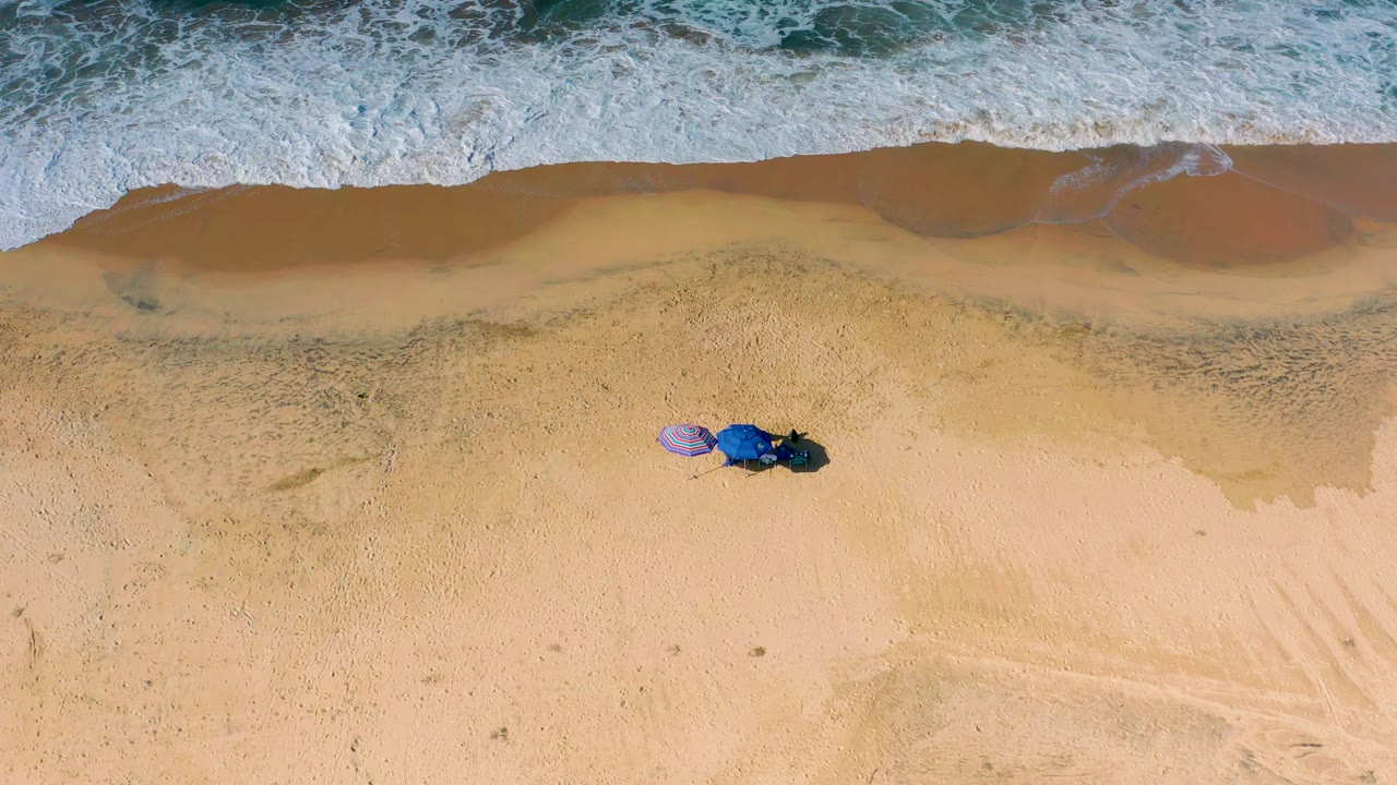 A straight-down Cerritos beach frame shows foamy water stretching along the top of the image while the purple and blue umbrellas sit on open sand with no visible people.