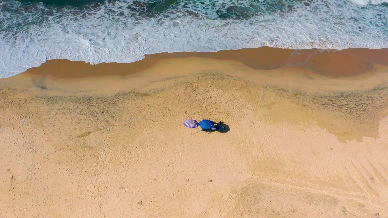 At Cerritos, Baja California Sur, a broad white shorebreak curls across the upper frame above two small umbrellas on tan sand, with no visible people.