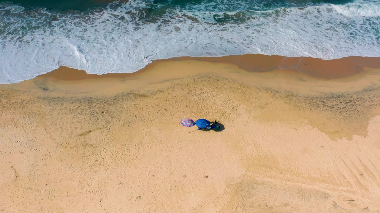 Aerial-looking frame over Cerritos beach with incoming foam occupying the top band, damp sand below it, and two umbrellas set back from the water with no visible people.