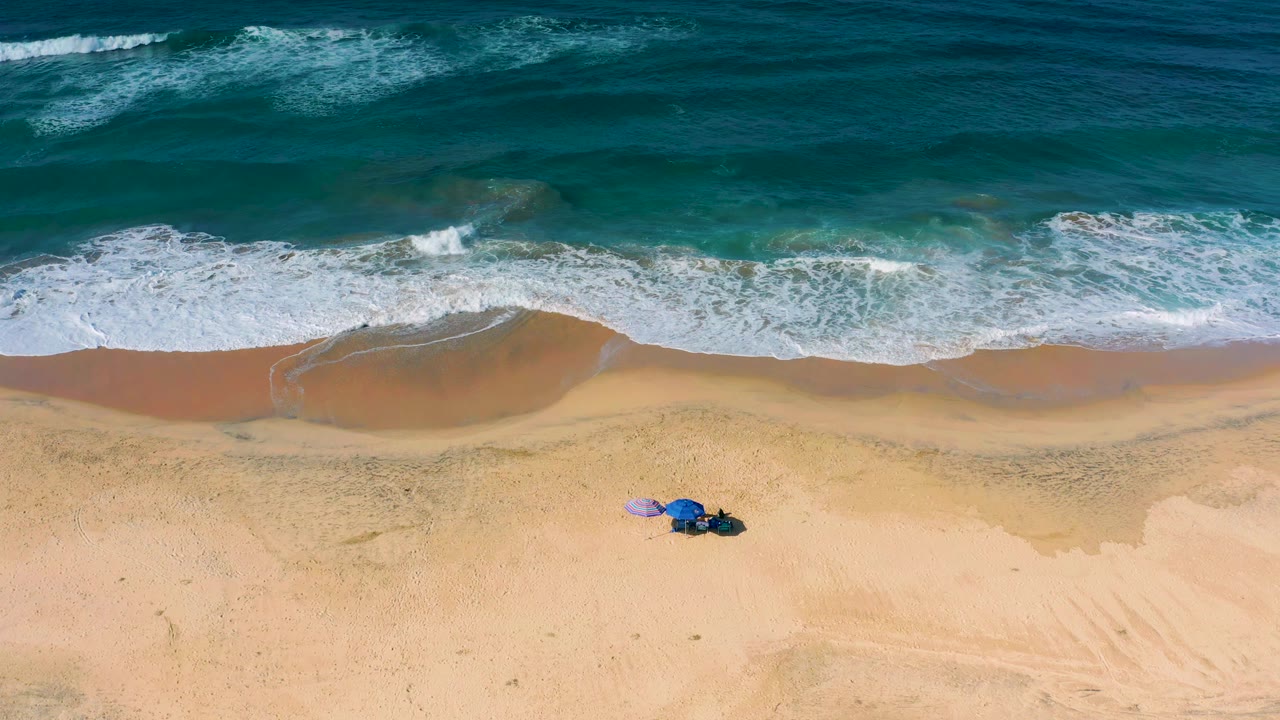 A later overhead frame shows the foam line spread more evenly across the Cerritos beach, with darker wet sand below the wave edge and no visible people.